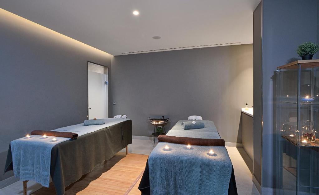 Mūsdienīgs spa telpa ar divām masāžas galdiem, kas izrotāti ar dvieļiem un svecēm, radot relaksējošu atmosfēru. Sienas ir krāsotas tumši pelēkā tonī, kas papildina telpas mierīgo noskaņu.