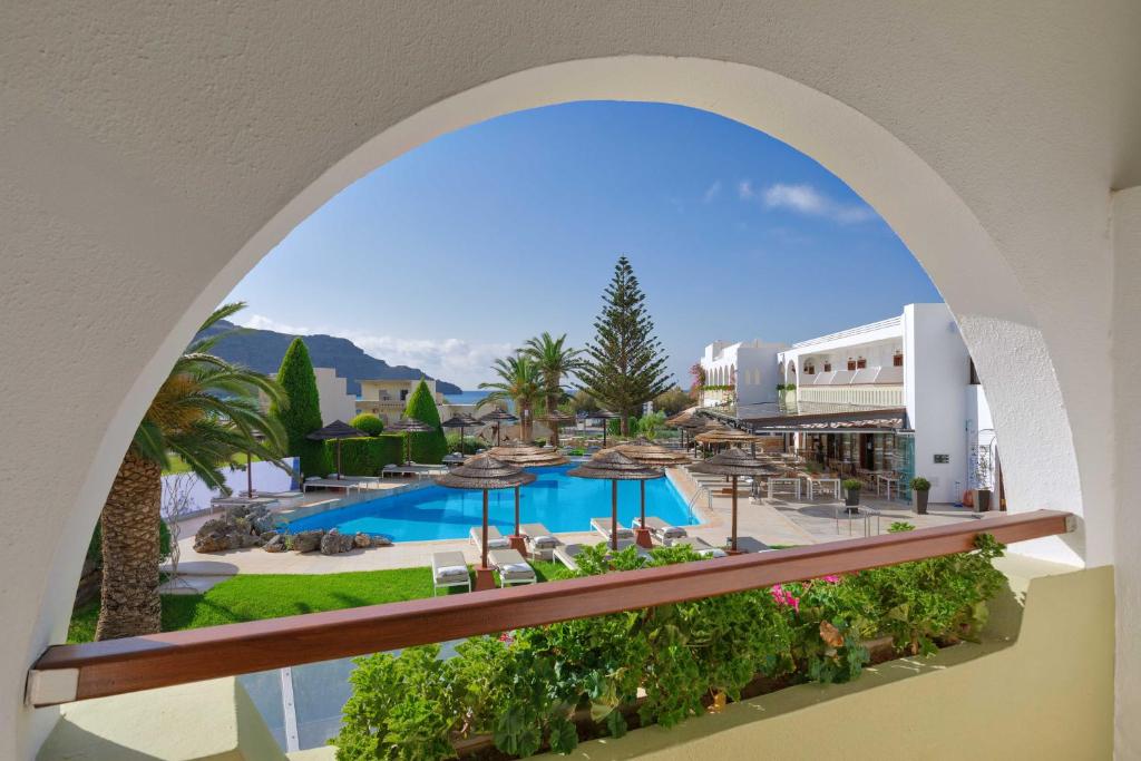 Skats no balkona uz viesnīcas baseinu, kurā ir sauļošanās krēsli un palmas, ar skaidru zilu debesi un kalniem fona.