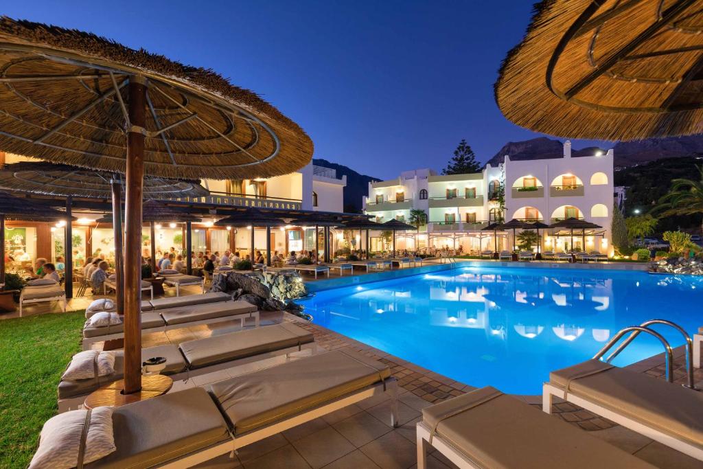 Luksusa viesnīca ar peldbaseinu un terasi, kas apgaismota vakara gaismā. Peldbaseins ir ieskauts ar sauļošanās krēsliem un palmu kokiem, radot relaksējošu atmosfēru.