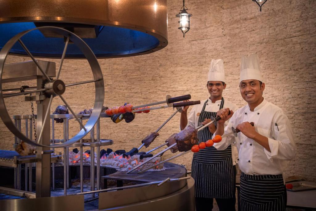 Šefpavāri pozē pie grila, kas rotē, turēdami gaļu un dārzeņus. Fons ir ar akmens tekstūru, radot siltu un viesmīlīgu atmosfēru.
