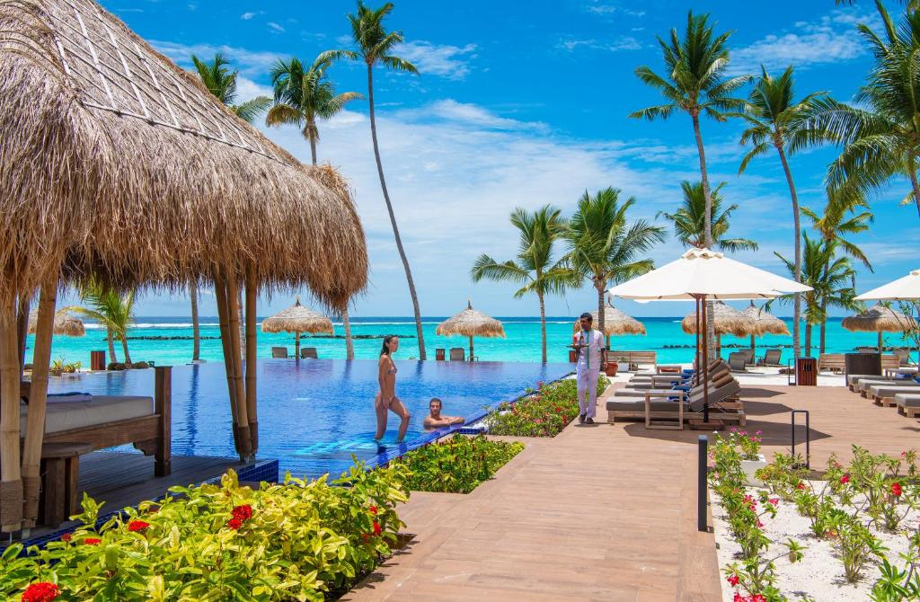 Tropu kūrorts ar bezgalības baseinu, palmām un saulainu debesi, kur cilvēki atpūšas pie ūdens. Ideāla vieta relaksācijai un atpūtai.