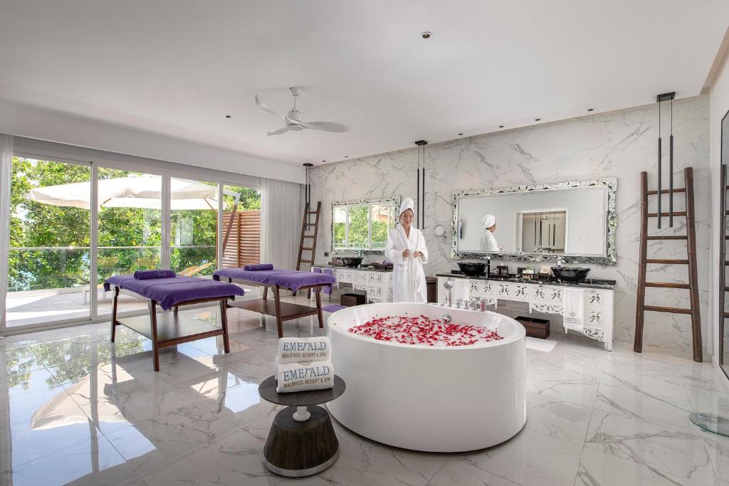 Luksusa spa telpa ar masāžas galdiem un baltu vannu, kas piepildīta ar ziedlapiņām. Telpa ir gaiša un moderna, ar lieliem logiem, kas sniedz skatu uz dabu. Spa darbinieks ir apģērbies baltā halātā un gaida viesus.