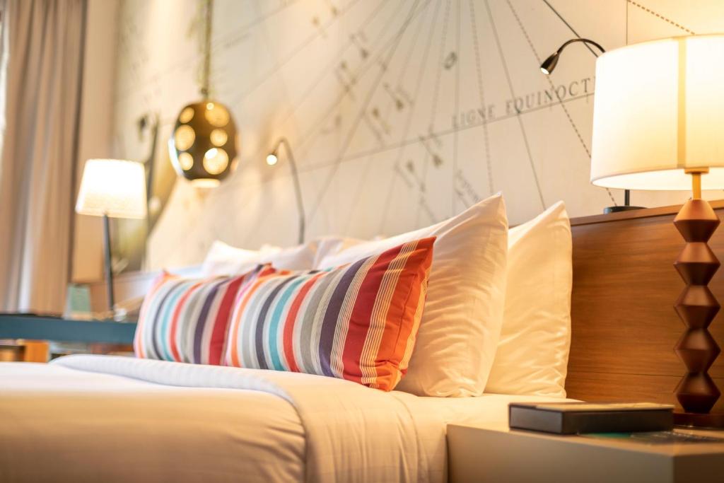 Mūsdienīgs guļamistabas interjers ar spilgtiem spilveniem un elegantu apgaismojumu, kas rada komfortablu atmosfēru. Fons ar grafiku papildina modernu dizainu.