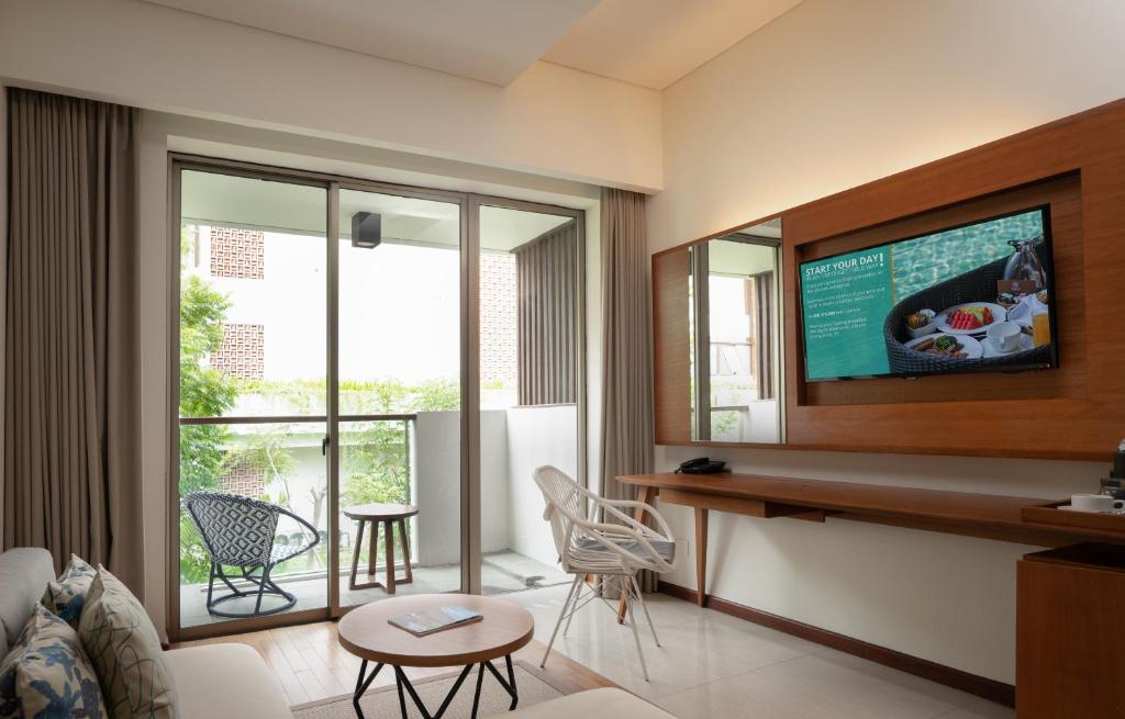 Mūsdienīga viesnīcas istaba ar lieliem logiem, kas ved uz balkonu, kur redzami zaļumi. Istabā ir eleganta mēbeļu kompozīcija, televizors un rakstāmgalds.