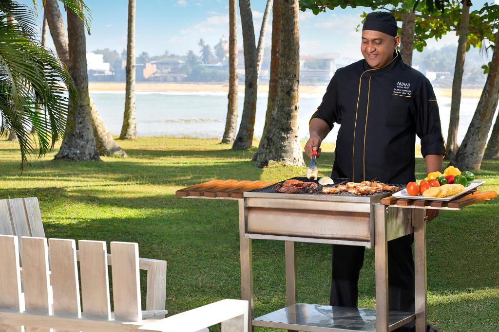 Grilējošs šefpavārs, kas gatavo gaļu un dārzeņus tropu ainavā pie pludmales. Apkārt ir zaļa zāle un palmām, radot relaksējošu atmosfēru.