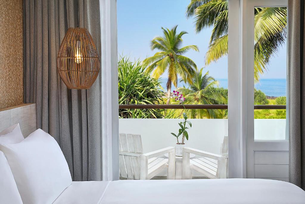 Mūsdienīga viesnīcas istaba ar skatu uz jūru, kurā ir gaisma no rotātas lampas un balta gulta. Terasē redzami palmas un sēdvietas.