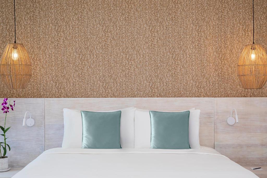 Mūsdienīgs guļamistabas interjers ar gaišu gultu, dekoratīvām spilveniem un ratiņu lampām, kas piekārti pie sienas ar teksturētu apdari. Florālais elements pie gultas piešķir dzīvīgumu.