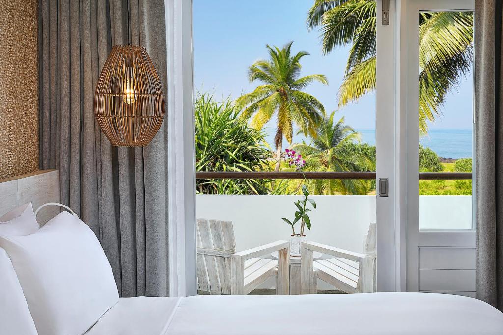 Mūsdienīga guļamistaba ar skatu uz jūru, kurā ir augu elementi un eleganta apgaismojuma lampa. Logā redzami palmu koki un atpūtas zona ar koka mēbelēm.