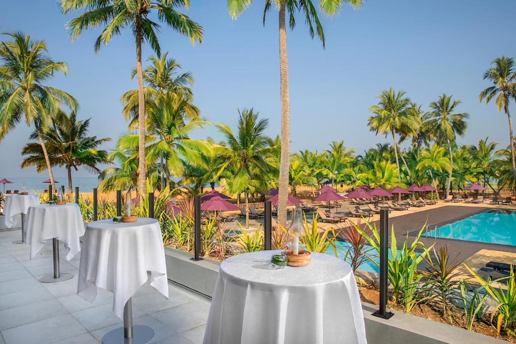 Tropu ainava ar palmām un peldbaseinu, kurā atrodas eleganti galdi ar baltām galdautām, rada relaksējošu atmosfēru. Fons ir skaists pludmales skats ar sauļošanās krēsliem un saulessargiem.