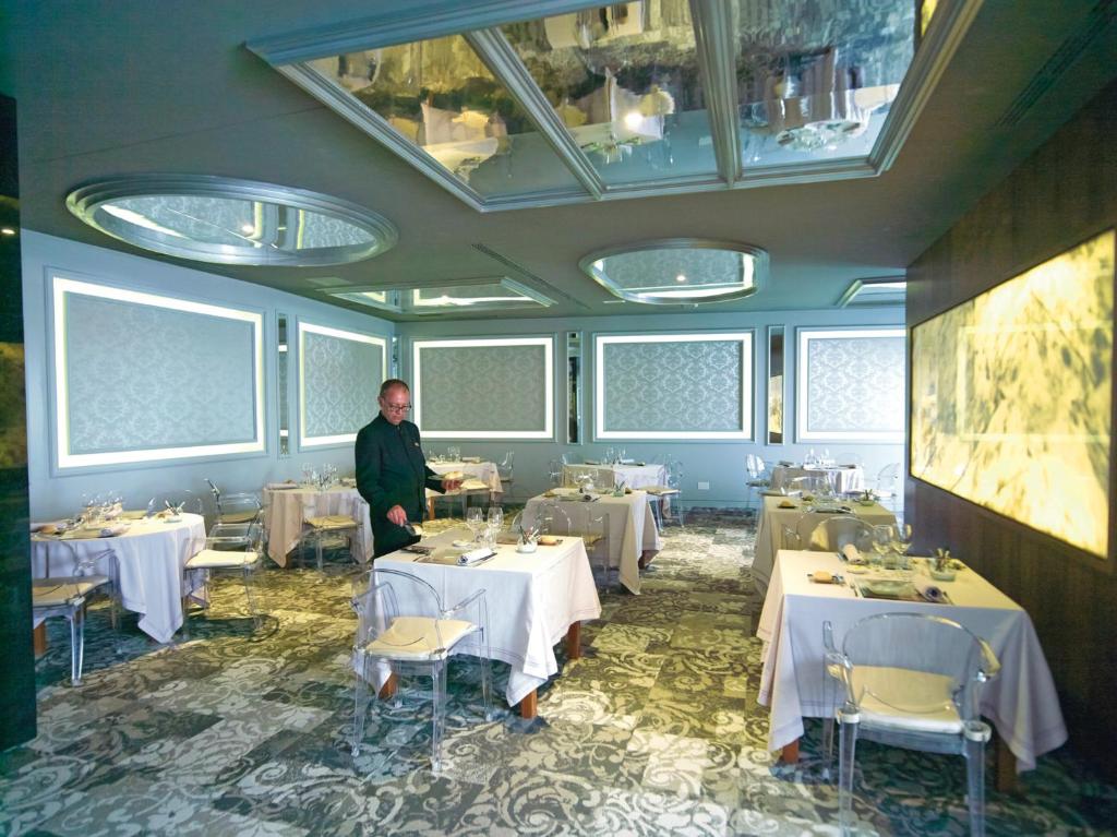 Eleganta restorāna iekārtojums ar stilīgām galdiem un gaismas elementiem. Gaisma atspoguļojas spoguļos, radot mūsdienīgu atmosfēru.