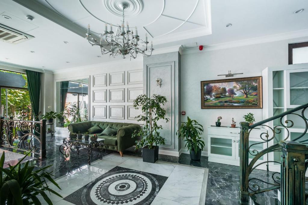 Mūsdienīgs viesistabas interjers ar zaļu dīvānu, greznu lustru un mākslas darbu pie sienas. Telpā ir daudz augi un lieli logi, kas nodrošina apgaismojumu.