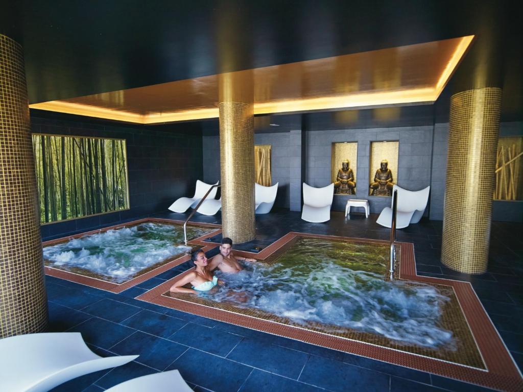 Mūsdienīgs spa ar džakuzi un relaksācijas zonu, kurā atpūšas pāris. Interjerā ir zeltainas kolonnas un dekoratīvi elementi, piemēram, statujas.