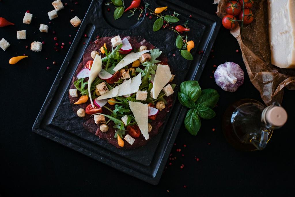 Svaiga salātu kompozīcija uz melna akmens trauka, dekorēta ar dārzeņiem, grauzdiņiem un sieru, kas papildināta ar garšaugiem un olīveļļu.