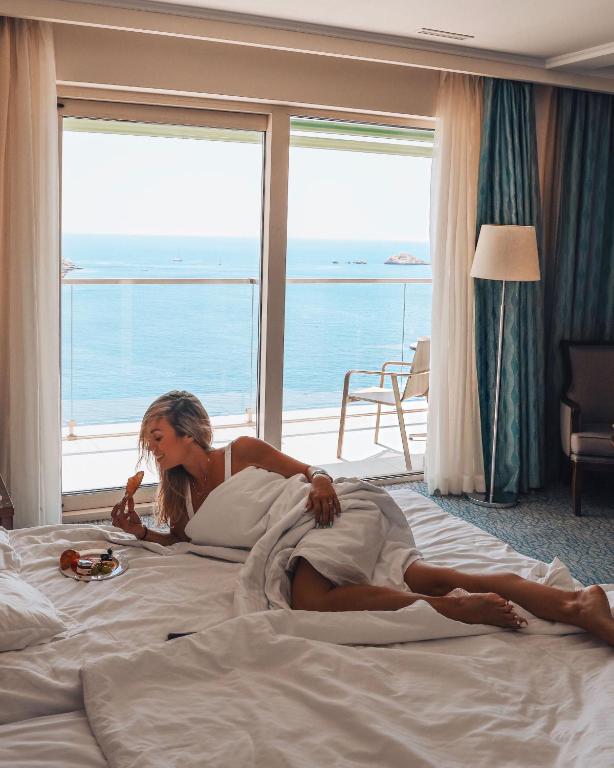 Sieviete atpūšas gultā, baudot brokastis, ar skatu uz jūru. Telpā ir gaiši aizkari un ērta mēbele.