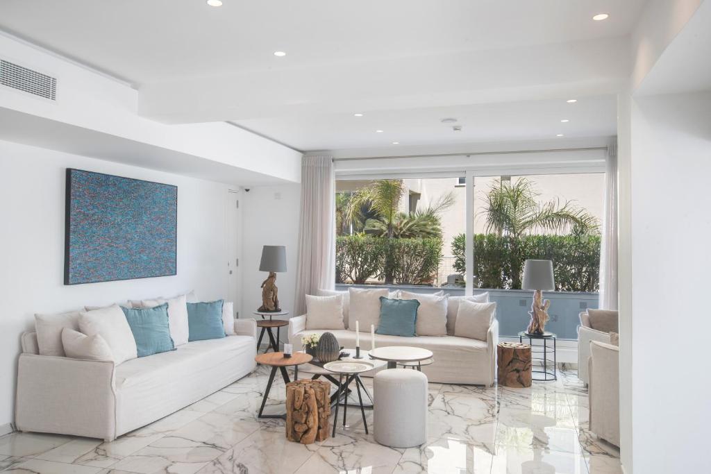 Mūsdienīgs dzīvojamās istabas interjers ar gaišām krāsām, elegantiem dīvāniem un dekoratīviem elementiem, kas rada relaksējošu atmosfēru. Lieli logi ļauj ieplūst dabiskajai gaismai un sniedz skatu uz zaļo dārzu.