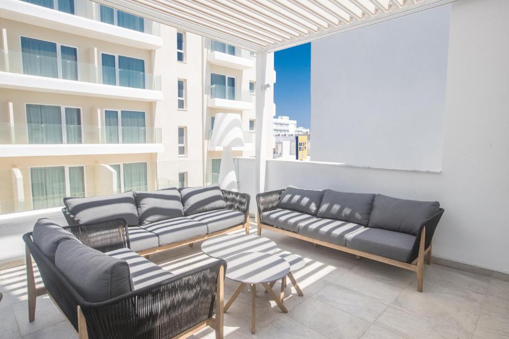 Mūsdienīgs āra atpūtas stūris ar pelēkiem dīvāniem un galdiņu, izgaismots ar saules stariem, fonā redzami koka paneļi un moderna arhitektūra.