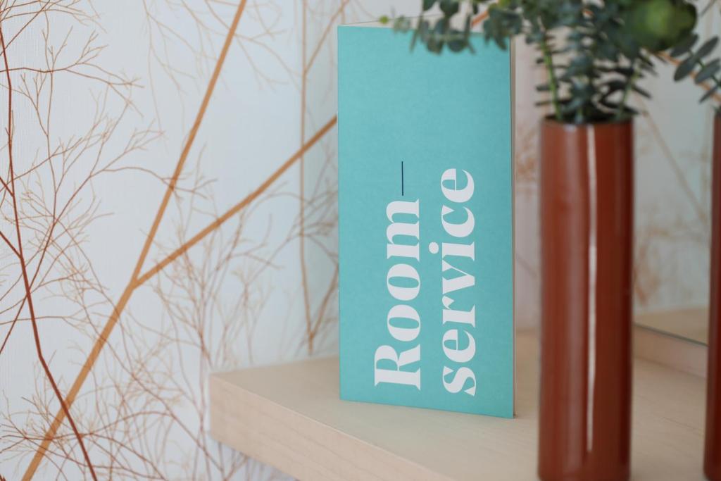 Zilā krāsā izstrādāta "Room service" brošūra uz koka virsmas, blakus brūnas krāsas vāzē ar zaļām augu zariem. Fons ir uzrakstīts ar dabīgiem motīviem.