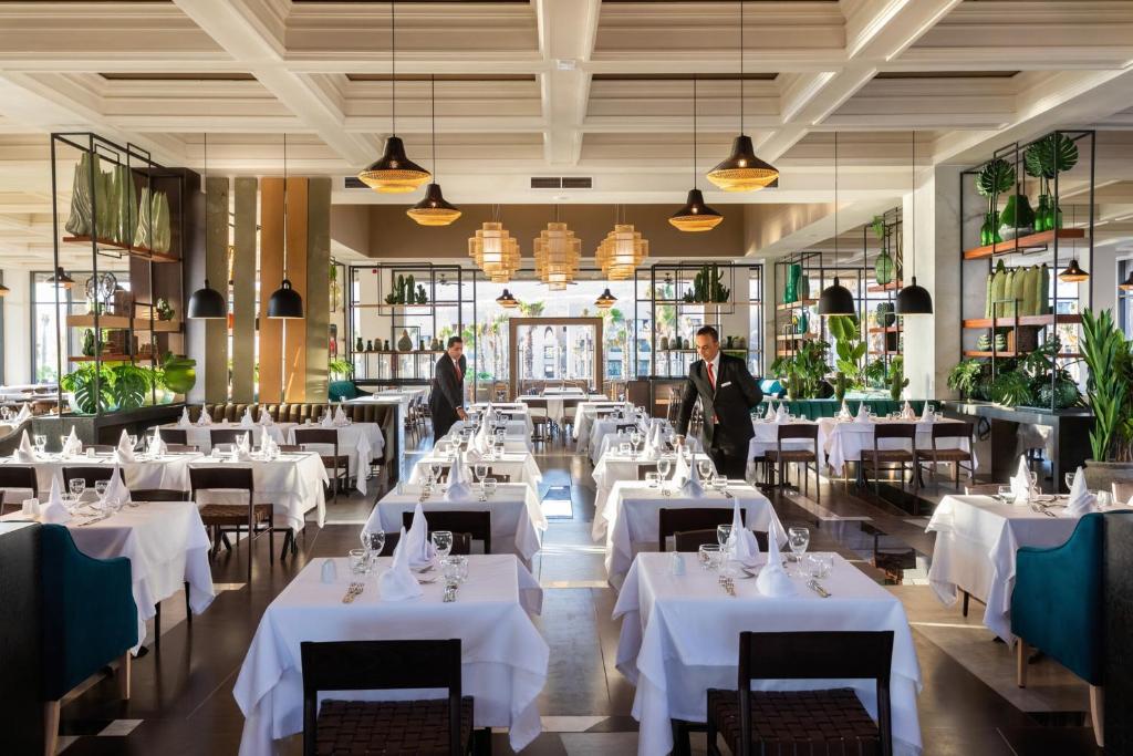 Moderns restorāns ar elegantiem galdautiem un zaļiem augiem, kas rada patīkamu atmosfēru. Apkalpojošais personāls sagatavo galdu, lai viesi varētu baudīt maltīti.
