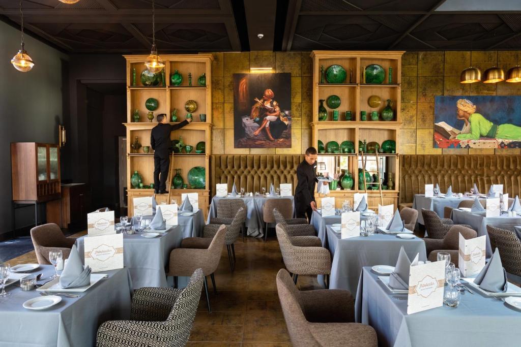 Mūsdienīgs restorāns ar elegantiem galdiem un zaļiem dekoratīviem priekšmetiem. Apkalpojošais personāls sakārto galdu un izkārtojumu, radot patīkamu atmosfēru.