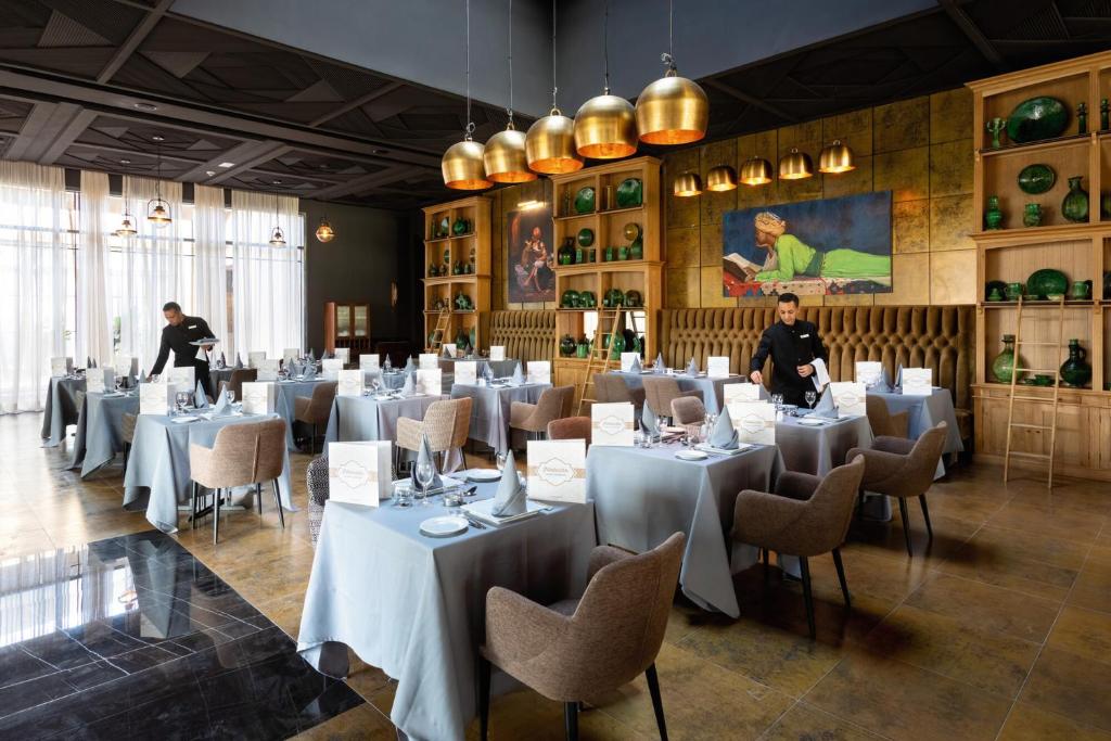 Mūsdienīgs restorāns ar eleganti noformētām galdiem un dekoratīvām interjera detaļām. Apkalpojošais personāls sagatavo zāli viesu ierašanās brīdim.