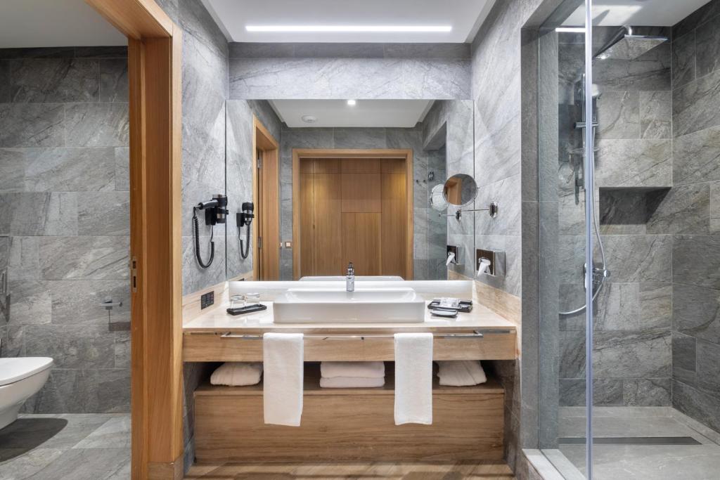 Moderns vannas istabas dizains ar akmens apdari, gaišām koka detaļām un stikla dušu. Eleganta un funkcionāla telpa ar plašu spoguli un vannas piederumiem.