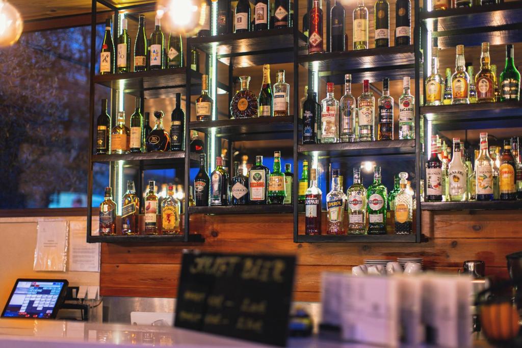 Bāra plaukts ar dažādām alkohola pudelēm, kas apgaismotas ar siltu gaismu. Priekšplānā redzams bāra galds un virtuves aprīkojums.