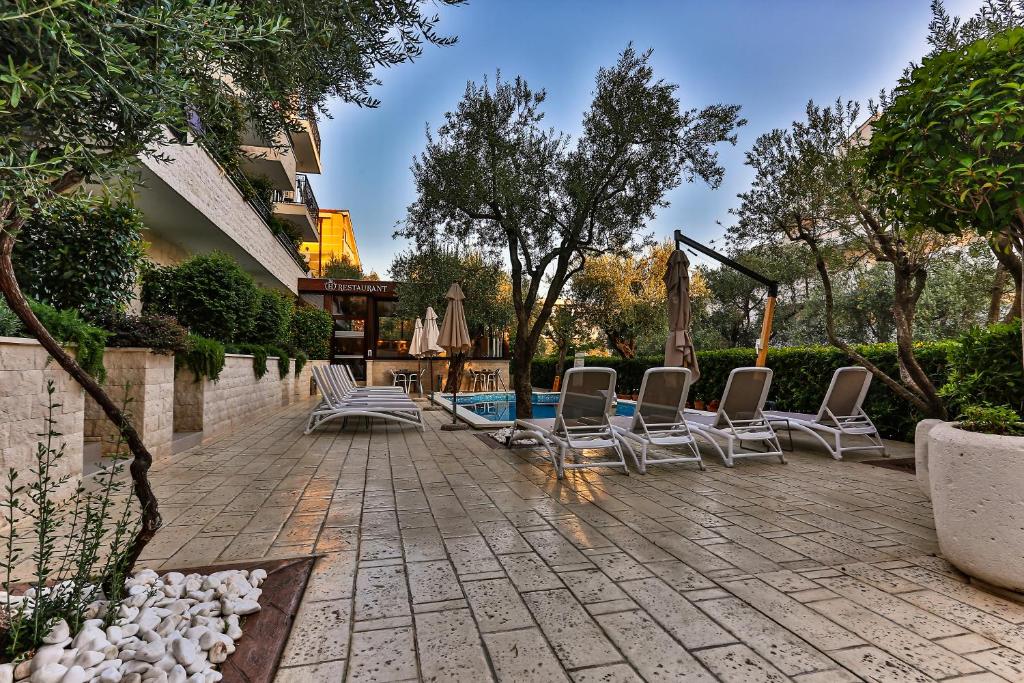 Relaksējoša atpūtas zona ar sauļošanās krēsliem, baseinu un zaļajiem kokiem. Vēl aiz tā redzams restorāns, kas piedāvā ēdienus svaigā gaisā.