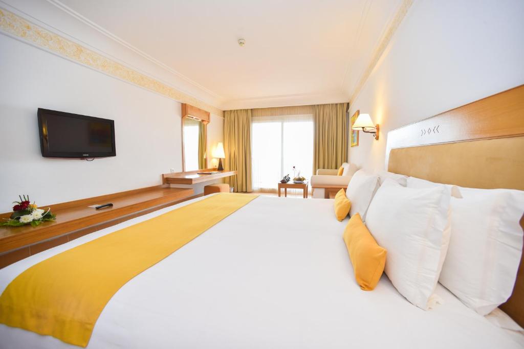 Mūsdienīga viesnīcas numura iekārtojums ar ērtu gultu un dzeltenu spilvenu, televizoru un logu, caur kuru ieplūst dabiska gaisma. Numurā ir arī dīvāns un galds, radot patīkamu atmosfēru atpūtai.