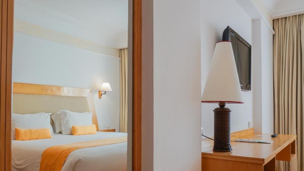 Mūsdienīga viesnīcas numura iekārtojums ar ērtu gultu, dekoratīvām spilveniem un gaismu, kas rada patīkamu atmosfēru. Numurā redzams arī rakstāmgalds un televizors, kas papildina komfortu.