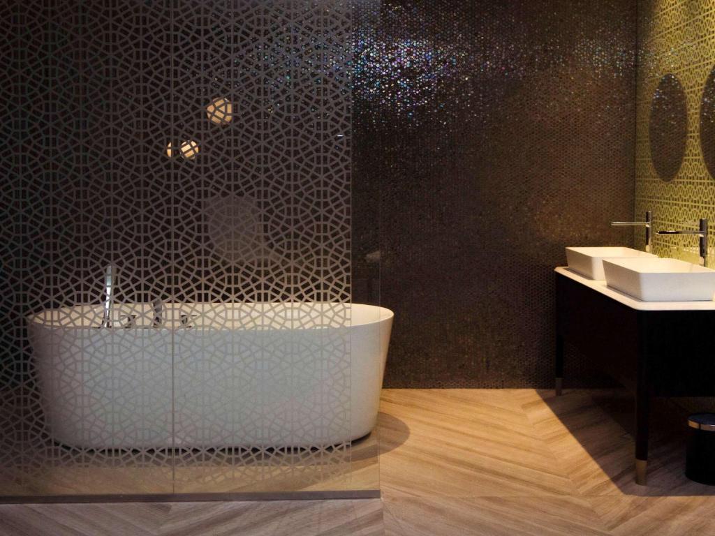 Mūsdienīgs vannas istabas interjers ar elegantu vannu un divām izlietnēm, akcentēts ar ģeometrisku rakstu uz sienas. Silta apgaismojuma atmosfēra un koka grīda piešķir telpai mājīgumu.