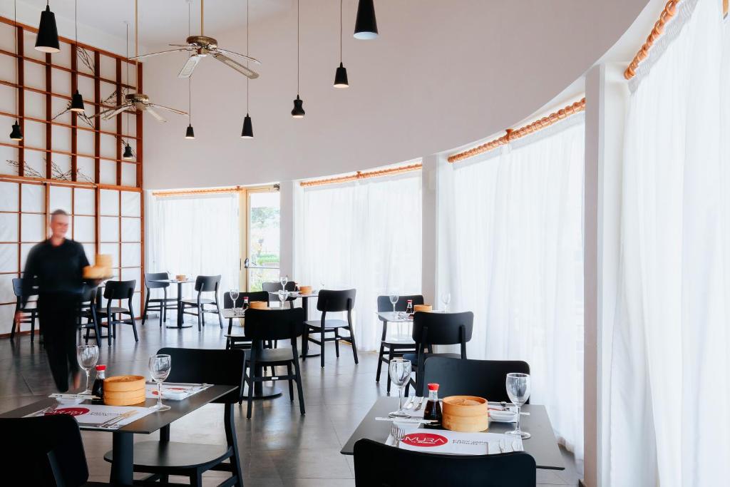Moderns restorāns ar elegantu interjeru, kurā redzami melni galdi un krēsli, kā arī gaismas piekari un caurspīdīgas aizkari. Apkārt ir klātas galdi, un viesmīlis pasniedz ēdienu.