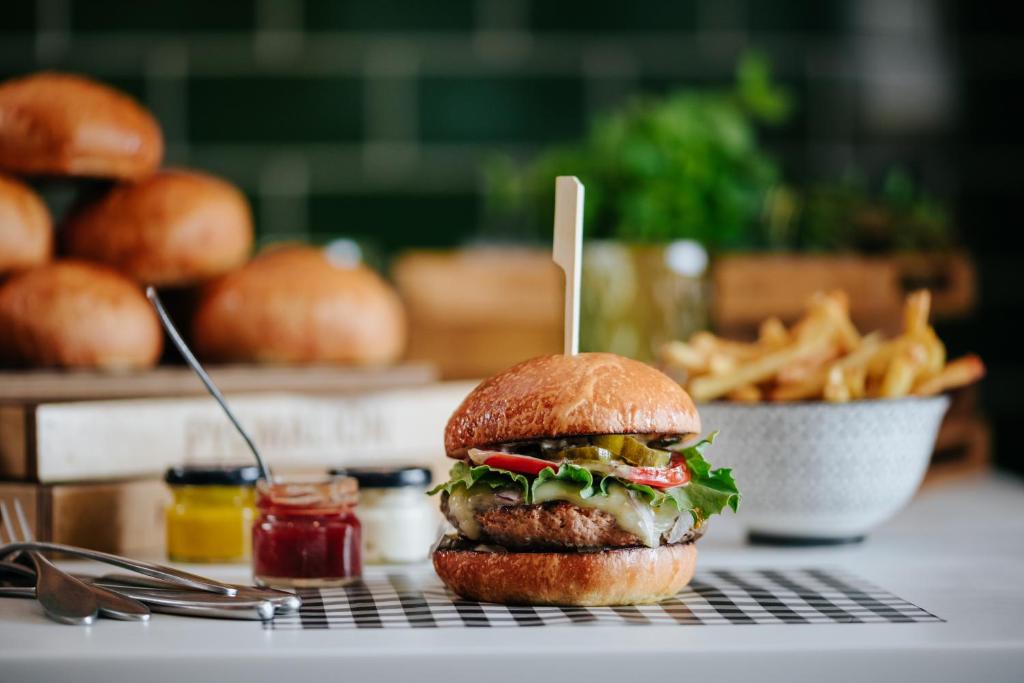 Svaigs burgeris ar salātiem, tomātiem un gurķiem, pasniegts uz melnbaltas pārbaudes formas. Fons ar maizēm un frī kartupeļiem papildina ēdiena izskatu.