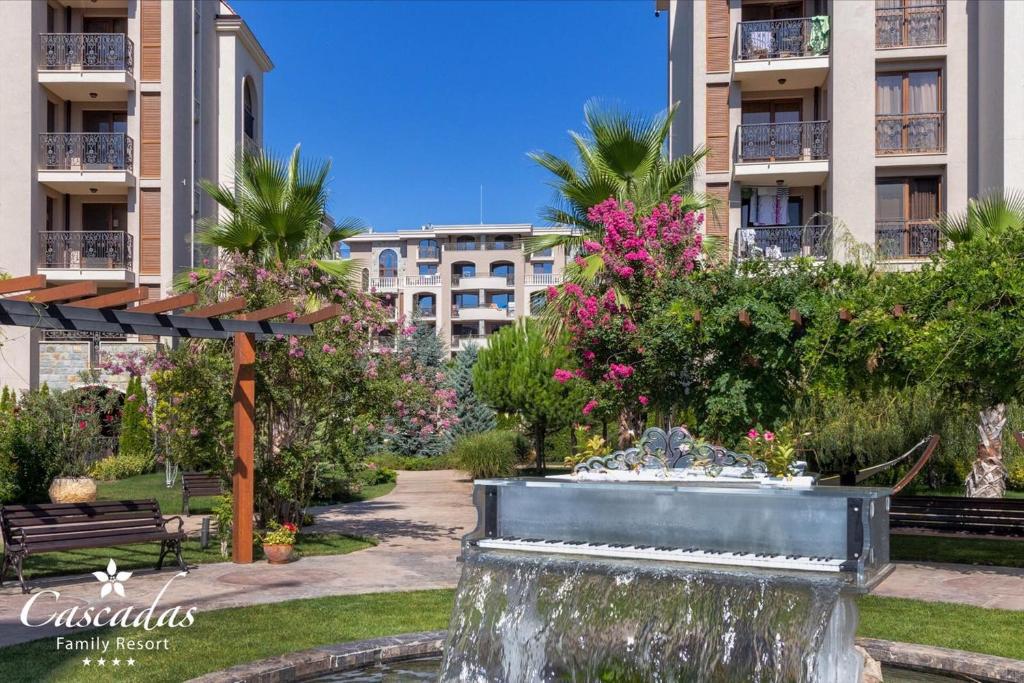 Dārza ainava ar strūklaku, ziediem un ēkām fona. Cascadas Family Resort logo apakšējā kreisajā stūrī.