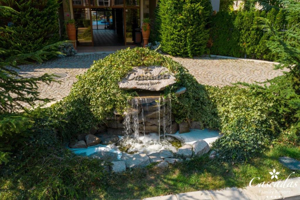 Dabīgā ūdens strūklaka ar akmeņiem un zaļumiem, kas rotā ieeju viesnīcā. Fonā redzama bruģēta aleja un viesnīcas ēka.