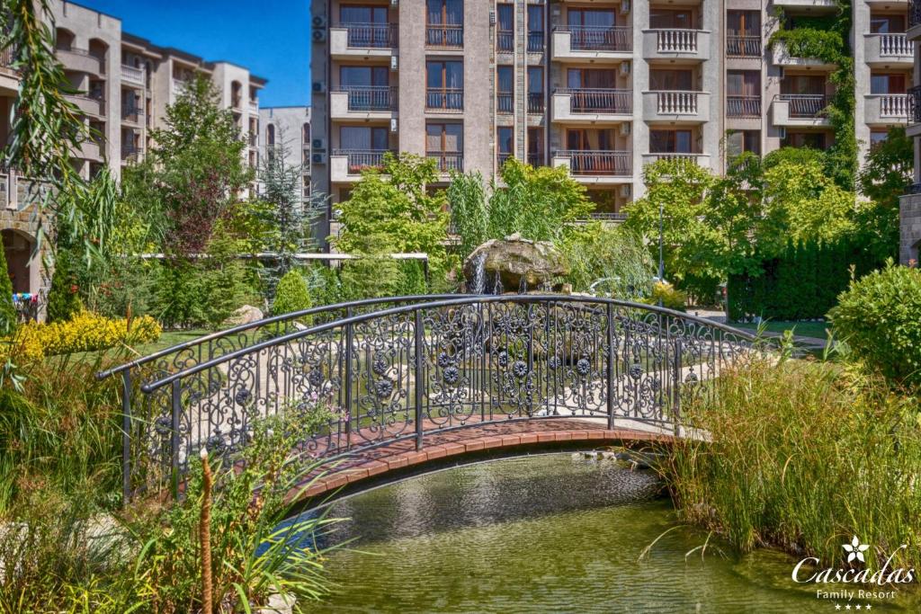 Dārza ainava ar metāla tiltiņu pār ūdeni, apkārt augošiem krūmiem un ziediem, un viesnīcas ēka fonā.