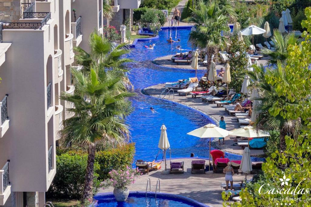 Peldbaseins ar zilu ūdeni un palmām, ap kuru atpūšas cilvēki uz sauļošanās krēsliem. Apkārt ir viesnīcu ēkas un zaļumi, radot atpūtas oāzi.