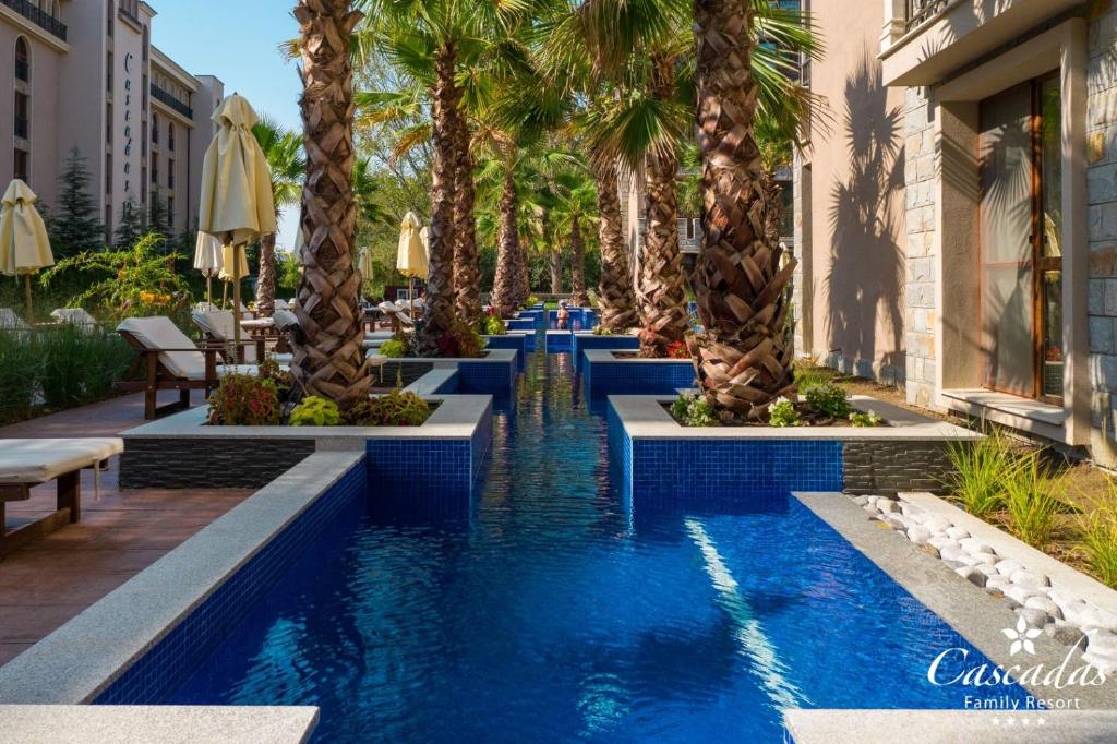 Peldbaseins ar palmām un saules krēslu rindu, radot relaksējošu atmosfēru pie Cascadas ģimenes kūrorta. Skats uz zilo ūdeni un zaļajiem augiem.