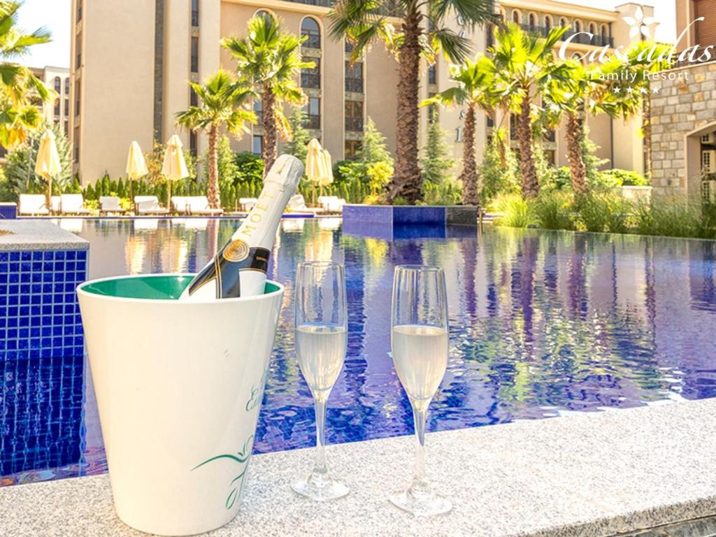Elegants šampanieša pudeles un divu flautu glāžu komplekts, kas izvietots pie baseina ar palmām un viesnīcas ēku fona. Ideāla vieta atpūtai un svinībām.