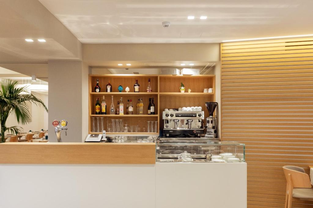 Mūsdienīga kafejnīca ar elegantu bāra zonu, kurā redzama kafijas automāts un dažādu dzērienu sortiments uz plauktiem. Interjers ir gaišs un viesmīlīgs, radot patīkamu atmosfēru.