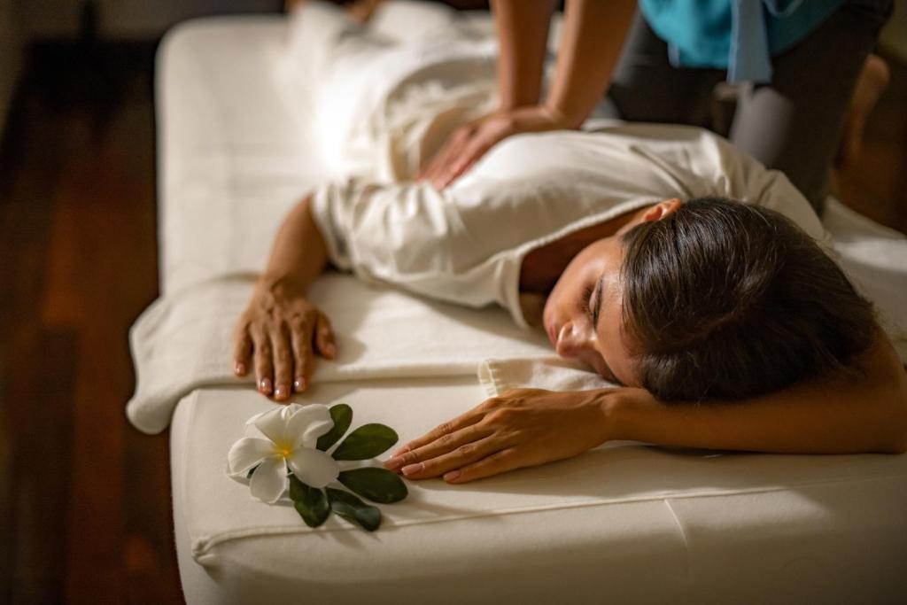 Masāžas sesija relaksējošā vidē, kur sieviete guļ uz masāžas galda, kamēr masieris veic masāžu, blakus novietota zieda kompozīcija.