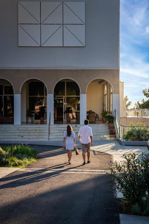 Pārī, kas iet pa celiņu uz moderna ēka ar arku logiem un augiem apkārt. Saules gaisma izgaismo ainavu, radot patīkamu atmosfēru.