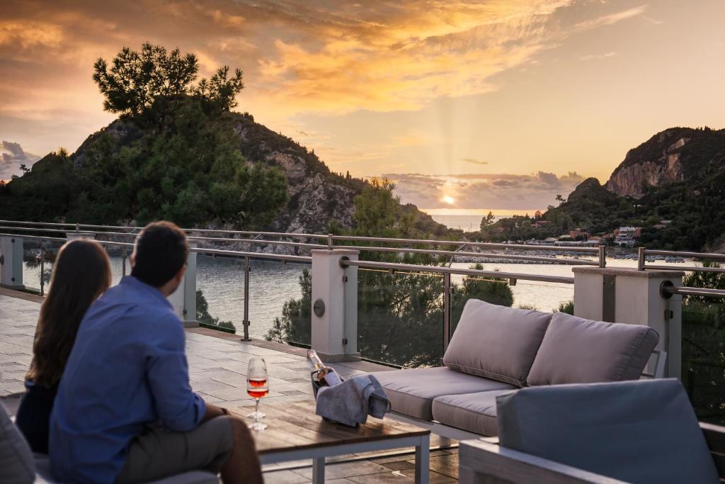 Romantisks skats uz saulrietu no terases, kur pāris piedalās mierīgā brīdī ar glāzi vīna rokās. Daba un jūra veido skaistu fonu, radot relaksējošu atmosfēru.