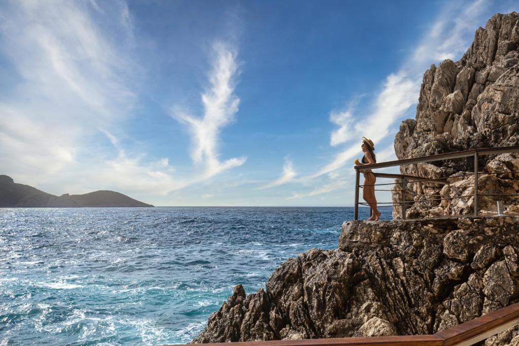 Sieviete stāv uz klinšu malas, skatoties uz plašo jūru un debesīm ar mākoņiem. Aiz viņas redzama kalnaina piekraste un mierīga jūras virsma.