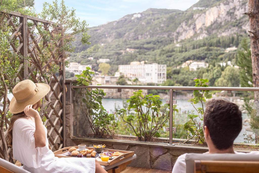 Relaksējoša ainava ar pāri, kas bauda brokastis uz terases, skatoties uz kalniem un ūdeni. Ideāla brīvdienu atmosfēra.