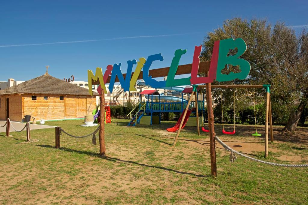 Bērnu rotaļu laukums ar krāsainu "Mini Club" zīmi, šūpuļtīkliem un slidkalniņu, kas atrodas saulainā vidē.