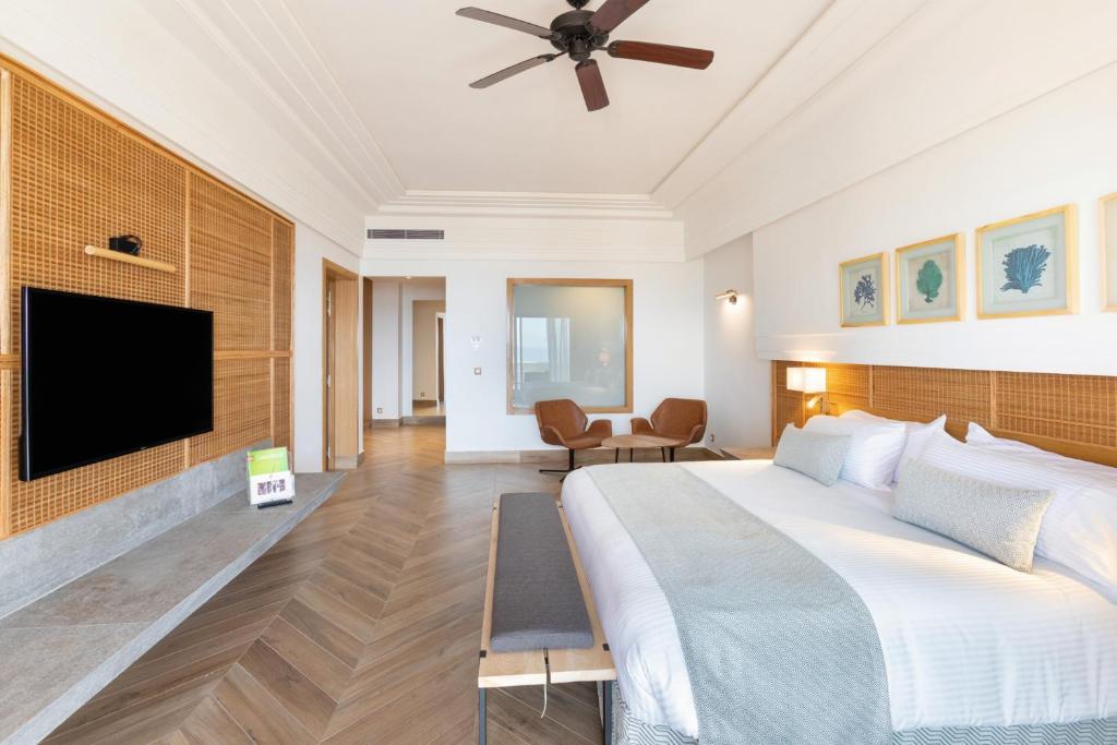 Mūsdienīga guļamistaba ar plašu gultu, koka grīdām un stilīgiem interjera elementiem, tostarp televizoru un mākslas darbiem uz sienas. Eleganta atmosfēra ar dabiskām krāsām un komfortablu sēdvietu.