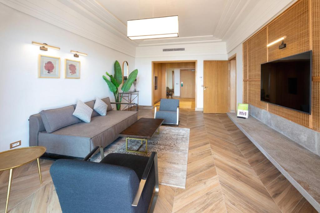 Mūsdienīgs viesistabas interjers ar pelēku dīvānu, melnu krēslu un koka grīdu, kurā ir liels televizors un dekoratīvas gleznas. Telpā ir daudz gaismas un zaļš augs, radot komfortablu atmosfēru.