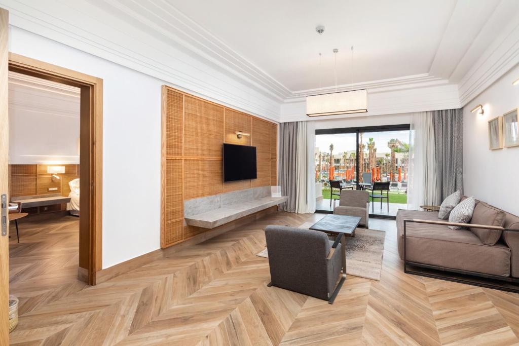 Mūsdienīgs viesnīcas numurs ar dzīvojamo zonu, koka grīdu un lieliem logiem, kas sniedz skatu uz āru. Interjers ir elegants un ērts, ar dīvānu, krēsliem un televizoru uz sienas.