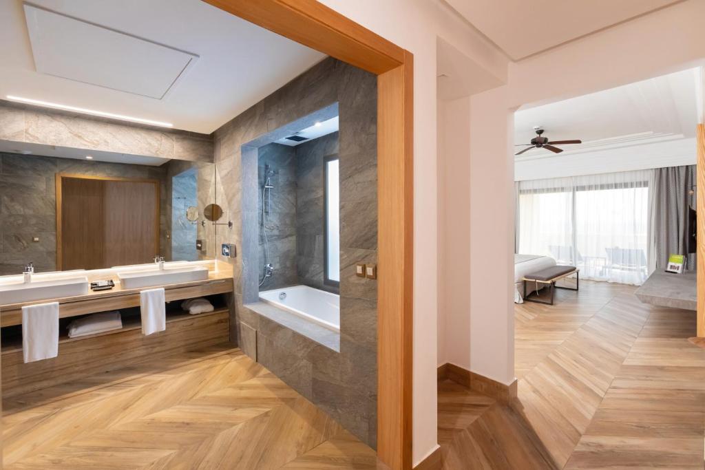 Mūsdienīgs vannas istabas interjers ar divām izlietnēm un vannu, kas nošķirts no guļamistabas ar koka grīdu un plašiem logiem, nodrošinot dabīgu apgaismojumu.