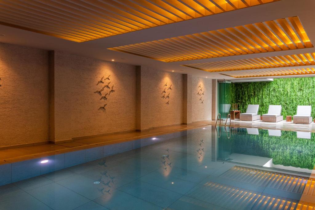 Mūsdienīgs iekštelpu peldbaseins ar relaksējošu atmosfēru, apgaismots ar siltām gaismām un dekorēts ar mākslinieciskām sienu rotājumiem. Peldbaseina mala ir apņemta ar ērtām atpūtas krēsliem un zaļu augu sienu.